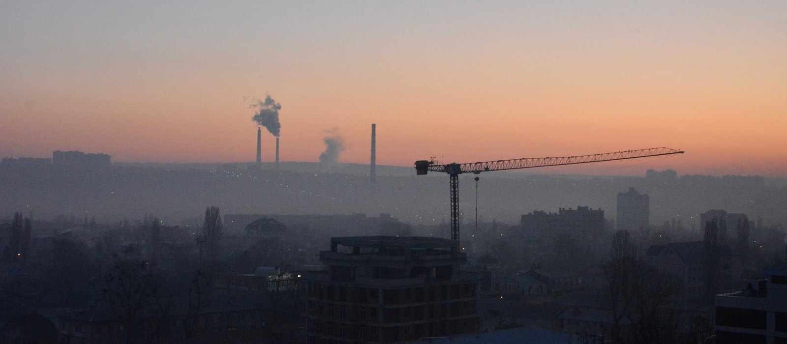 Chișinăul în zori, cu o macara deasupra unui bloc în construcție și coșurile unei centrale termice fumegând în ceață