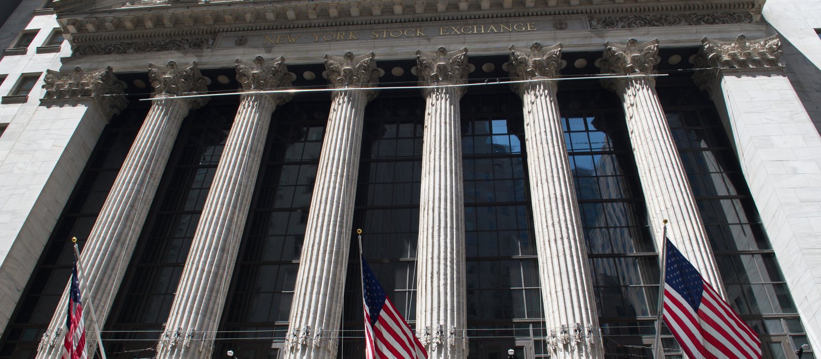 Fațada cu șase coloane corintice a clădirii New York Stock Exchange, văzută de jos, cu steaguri americane la baza coloanelor